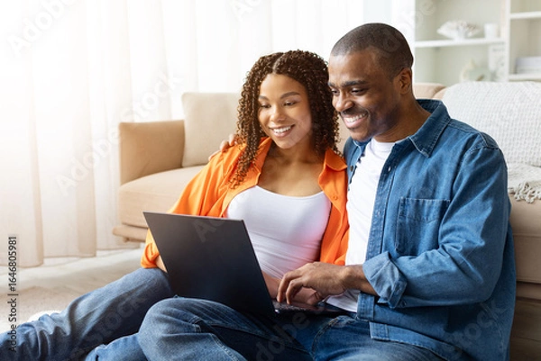 Fototapeta Smiling young African American couple sitting on floor with laptop at home, browsing together and enjoying time in casual relaxed setting, copy space