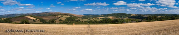 Fototapeta Late-summer panorama of a harvested field with a single straw bale, rolling patchwork hills and scattered clouds in Shropshire, UK.