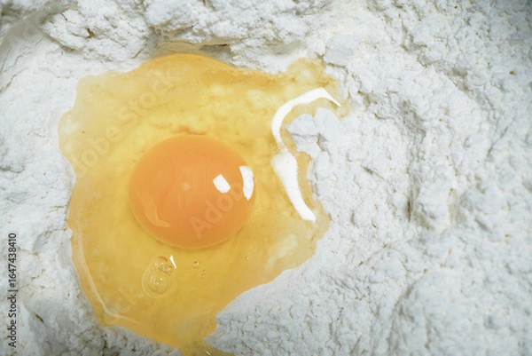 Fototapeta Cracked egg rests in mound of white flour, ready for baking. yolk is vibrant and egg white is slightly spread, creating contrast with flour. This setup is perfect for making dough or batter