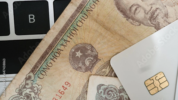 Fototapeta Close up image of Vietnamese currency notes partially covering laptop keyboard, with credit card placed on top, suggesting themes of finance and technology