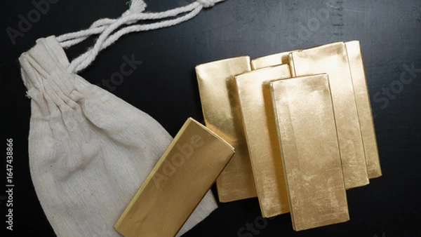 Fototapeta Gold bars arranged on dark surface next to white cloth bag, creating sense of wealth and luxury. shiny metal reflects light, emphasizing its value and allure