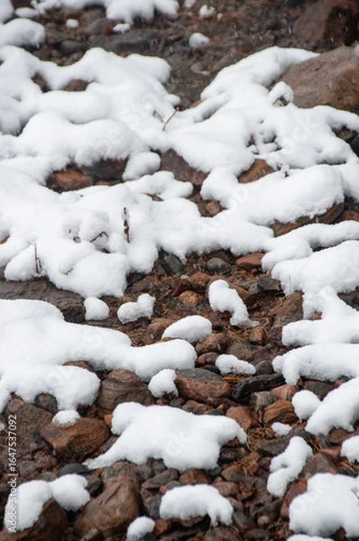 Fototapeta A detailed vertical shot of a ground covered in brown and grey rocks, with soft, white patches of fresh snow creating an interesting texture and pattern.