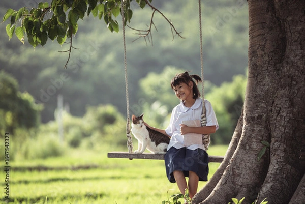 Fototapeta Young girl sitting on swing under tree with cat in peaceful rural landscape, enjoying nature and companionship in summer sunlight, serene atmosphere and joyful moment