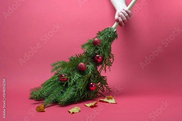 Fototapeta Hand sweeps autumn leaves with a festive evergreen broom adorned with red baubles, signaling cheerful holiday cleanup and fresh start.