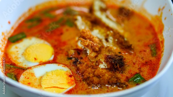 Fototapeta Close-up of a vibrant spicy curry noodle soup featuring crispy fried chicken hard-boiled eggs and fresh green vegetables in rich red-orange broth.