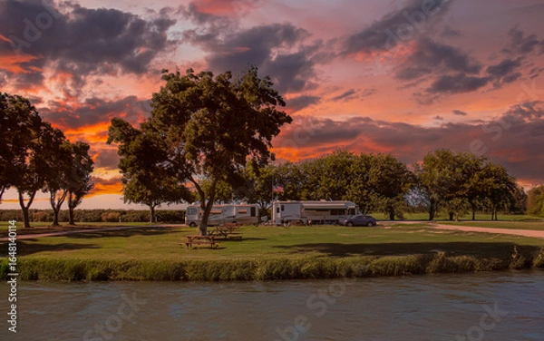 Obraz RVs are parked near a serene lakeside surrounded by lush trees, under a vibrant sunset sky. The calm water reflects the stunning colors of the sky, creating a tranquil scene.