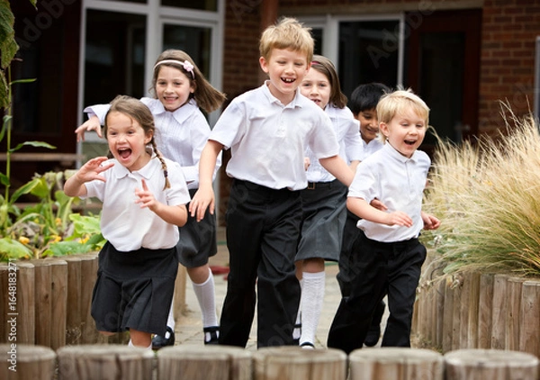 Fototapeta Cheerful primary students running outdoors. A group of happy children in school uniforms laughing and playing in a school playground during break time.
