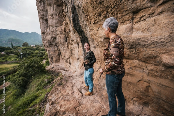 Fototapeta Mother and daughter on a hike through the countryside where they found cave paintings on a cliff, dressed in hiking clothes.