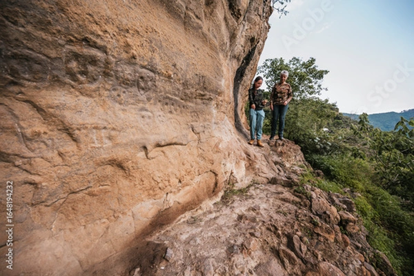 Fototapeta Mother and daughter on a hike through the countryside where they found cave paintings on a cliff, dressed in hiking clothes.