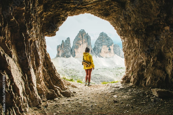 Fototapeta Explorer in Yellow Jacket Framed by Cave Opening with View of Tre Cime di Lavaredo Peaks
