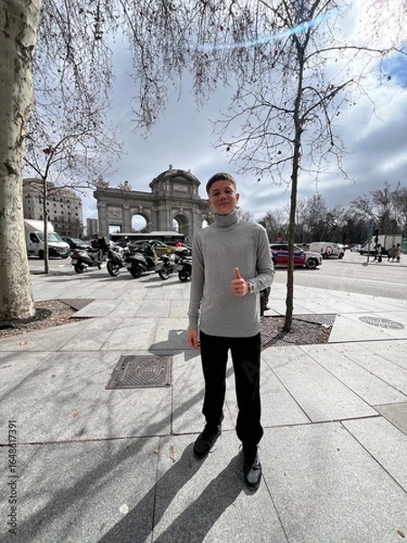 Fototapeta City Explorer's Stroll: A young person strolls confidently along a city sidewalk, their thumb raised in a gesture of optimism, with a majestic arch providing a stunning backdrop.