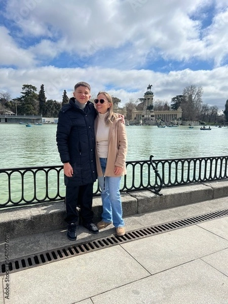 Fototapeta Couple's Leisure: A couple, immersed in their leisure, poses beside a tranquil lake, with architectural marvels adding an air of grace to this picturesque outdoor scene.