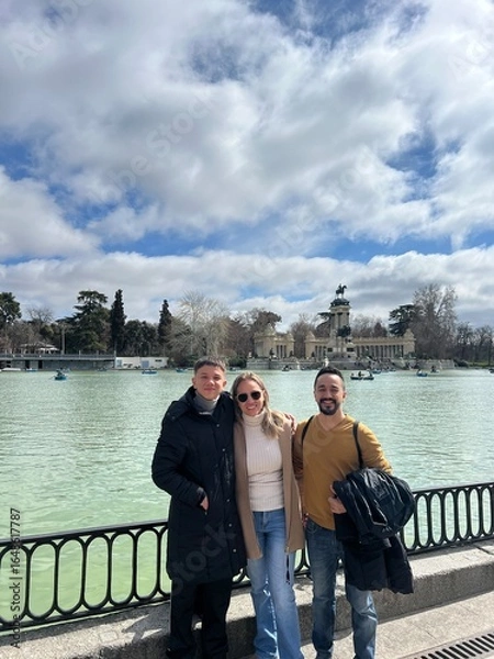 Fototapeta Friends by the Lake: Three friends pose happily near a serene lake with an ancient building as a backdrop, enjoying a leisurely day out under a vibrant blue sky with fluffy clouds.