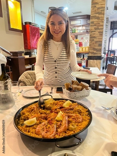 Fototapeta Delighted Dining Moment: A smiling individual presents a delectable seafood paella at a table, showcasing culinary skill and joy.