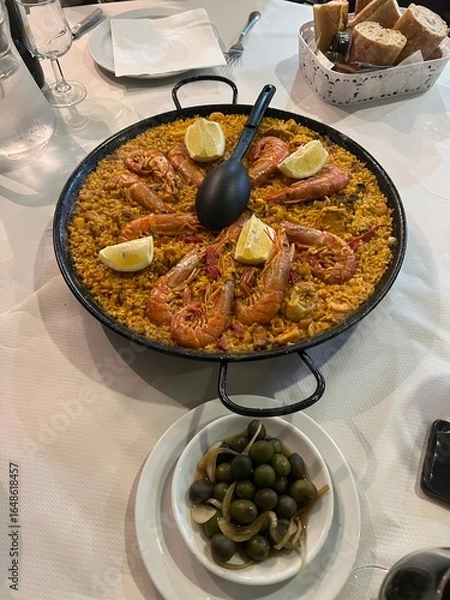 Obraz Exquisite Paella Feast: A vibrant paella dish, brimming with succulent shrimp and zesty lemon wedges, served alongside olives and crusty bread, offering a tantalizing culinary experience.