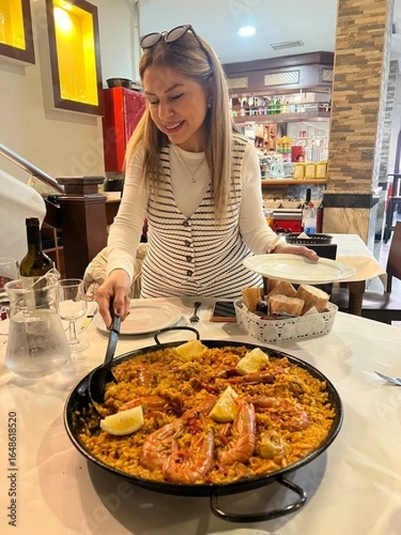 Fototapeta Exquisite Paella Presentation: A woman delicately serves a vibrant paella dish, featuring succulent shrimp and zesty lemon wedges, within a classic dining establishment.