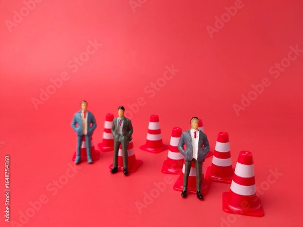 Fototapeta The image features a three miniature figurine of a person standing on a traffic cone, with other traffic cones on a red background