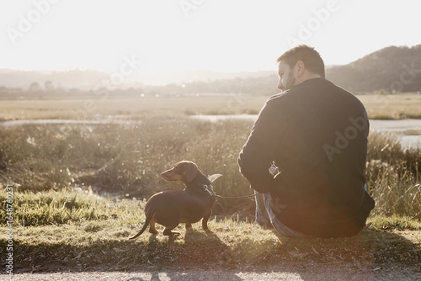 Fototapeta Man in black long sleeved shirt and blue jeans sitting out in nature next to a lake with a brown dachshund in a harness and on leash engaging with his pet dog. Sun flare. Lake and grass
