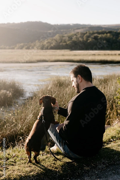 Fototapeta Man in black long sleeved shirt and blue jeans sitting out in nature next to a lake with a brown dachshund in a harness and on leash engaging with his pet dog. Sun flare. Lake and grass