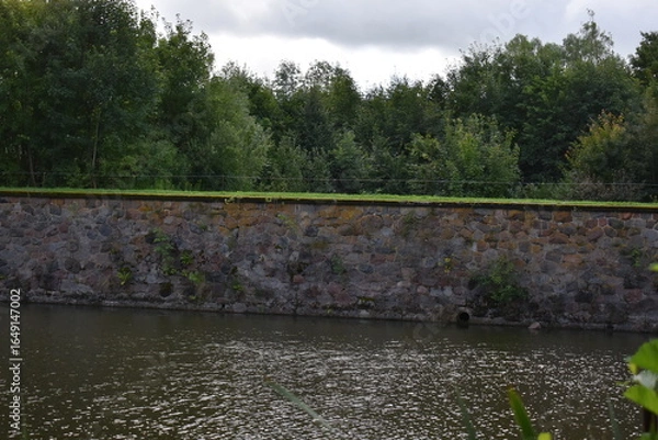Obraz Stone wall along river surrounded by trees under cloudy sky  
