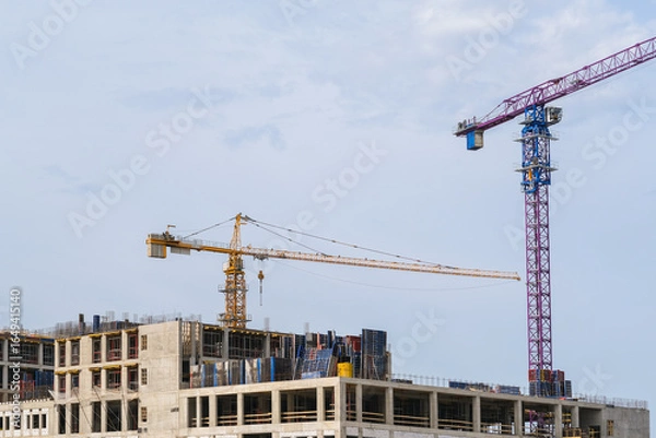 Fototapeta Two tall construction cranes overlooking the concrete structures of a building site under a clear sky. Real estate development.