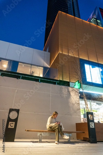 Fototapeta Urban Contemplation: A person sits on a bench, engrossed in their phone, amidst a backdrop of modern architectural marvels and twilight hues. A scene of serene solitude in the urban landscape.