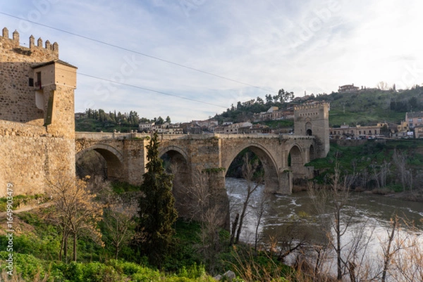 Fototapeta Toledo Bridge: Spain's historic San Martín Bridge, gracefully spanning the Tagus River, is steeped in history and culture, with medieval stone arches, towers, and a timeless design under a sunny sky
