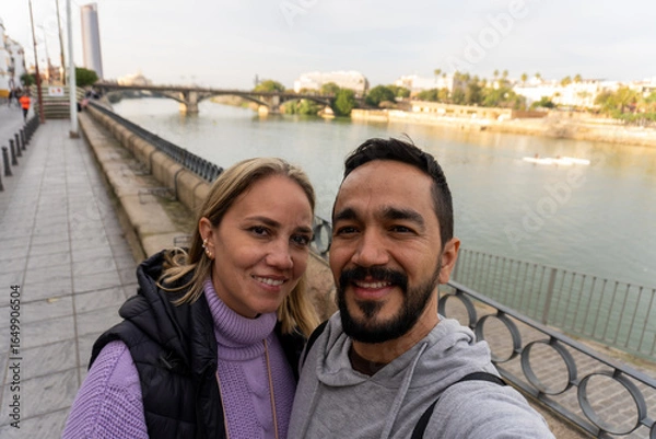 Fototapeta Selfie on a scenic bridge in Seville, Spain: a couple poses on a bridge in front of the camera, with a beautiful river and cityscape in the background.