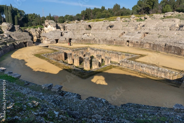 Obraz Ancient Roman Amphitheater itálica: A grand stone amphitheater stands proudly against a backdrop of verdant trees, the arena's walls weathered by time and history.