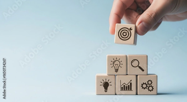 Fototapeta Wooden cubes with business icons, hand placing a target cube on top, light blue background, showcasing goal achievement and strategic planning