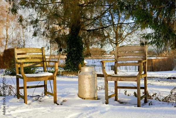 Fototapeta Two empty wooden chairs and a vintage milk can sit in a tranquil, snowy garden during a sunny winter day, evoking a sense of peace and nostalgia.
