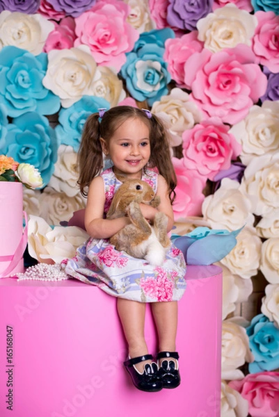 Fototapeta Happy laughing little girl playing with a baby rabbit, hugging her real bunny pet and learning to take care of an animal. Child on a floral background
