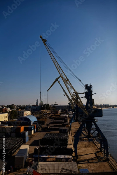 Fototapeta Large Gantry Crane at Industrial Waterfront with City Skyline. A striking yellow crane stands tall on a pier, with a vast expanse of water and distant cranes, highlighting a bustling port scene bathed