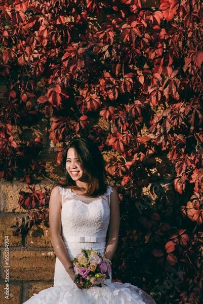 Fototapeta Bride stands by the wall with red leaves
