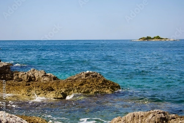 Fototapeta Landscape with sea and coastal rocks. The water shimmers with shades of blue and turquoise, and a small island with green vegetation is visible on the horizon.