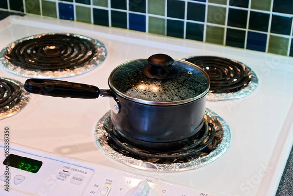 Fototapeta Stainless steel saucepan boils vigorously on modern electric cooktop, glass lid revealing steam as food cooks inside. White stove shows digital timer display set to 7:22, with green and blue mosaic