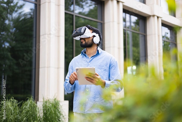 Obraz A man wearing a VR headset and headphones, holding a tablet in front of a building.
