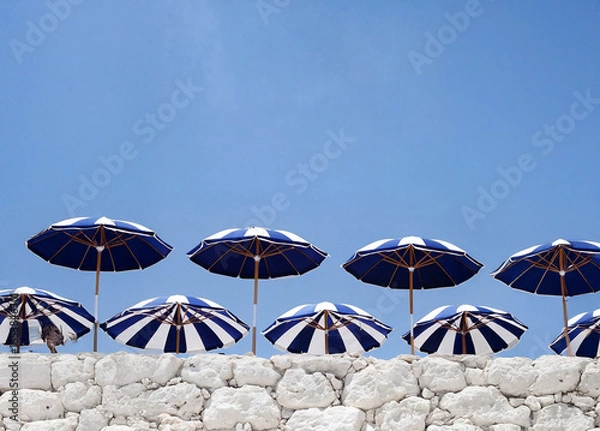 Fototapeta A series of blue and white striped umbrellas arranged on a white stone wall against a clear blue sky.