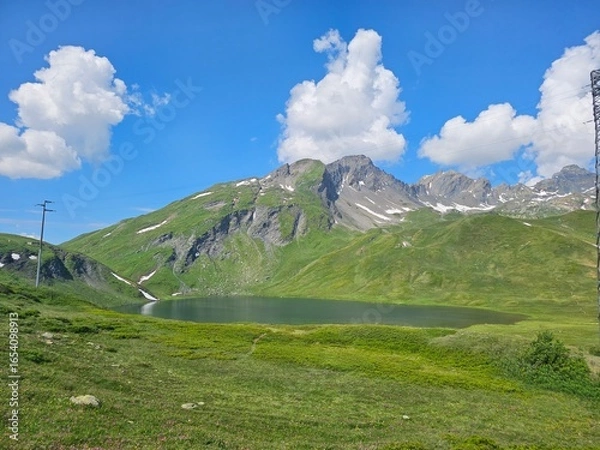 Fototapeta La Thuile, Val D'Aosta, July 02 2025: Panoramic view to the alps, beautiful green landscape reflected in a small natural lake. Beautiful blue sky with some clouds. Mountains with a little snow. Aerial