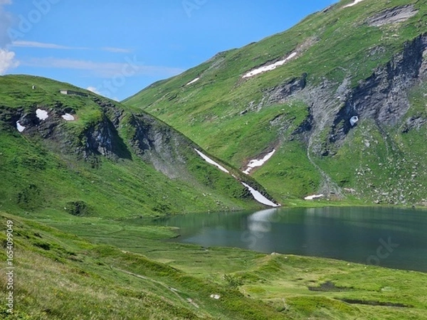 Fototapeta La Thuile, Val D'Aosta, July 02 2025: Panoramic view to the alps, beautiful green landscape reflected in a small natural lake. Beautiful blue sky with some clouds. Mountains with a little snow. Aerial