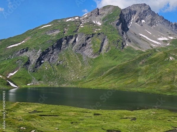 Fototapeta La Thuile, Val D'Aosta, July 02 2025: Panoramic view to the alps, beautiful green landscape reflected in a small natural lake. Beautiful blue sky with some clouds. Mountains with a little snow. Aerial