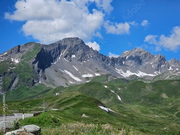 Fototapeta La Thuile, Val D'Aosta, July 02 2025: Panoramic view to the alps, beautiful green landscape reflected in a small natural lake. Beautiful blue sky with some clouds. Mountains with a little snow. Aerial