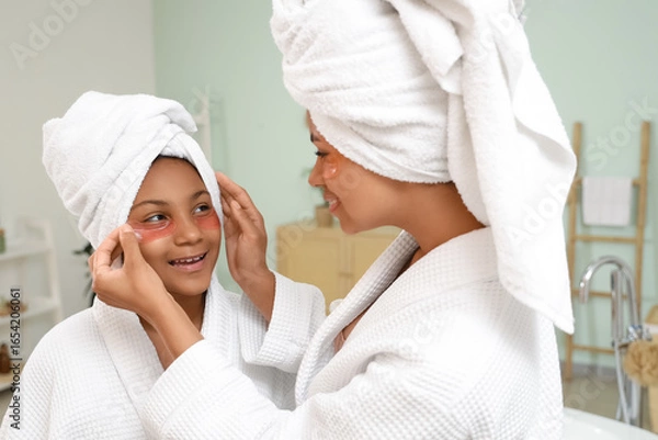 Obraz Happy young African-American mother and her daughter in bathrobes applying under-eye patches at home