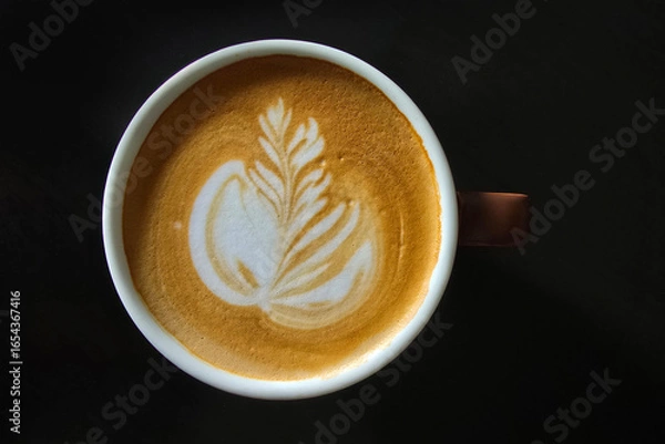 Fototapeta A close-up top view of a freshly brewed latte with intricate leaf-pattern latte art served in a ceramic cup on black background.
