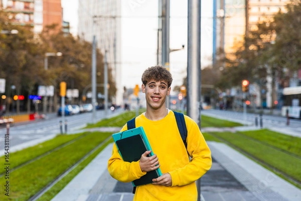 Obraz Smiling college student holding books and walking in city center