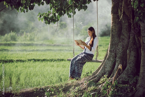 Fototapeta Beautiful Asian woman in traditional dress enjoys reading a book on a swing while enjoying nature and fresh air in a rice field in Isan, Thailand.