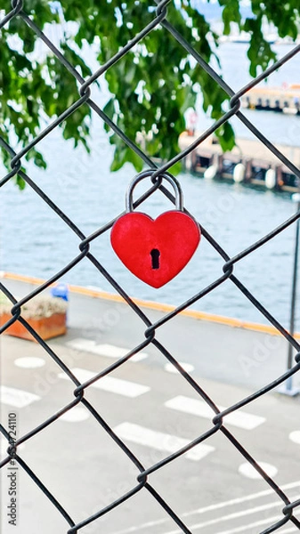Obraz A heart-shaped padlock attached to a chain link fence with an urban cityscape visible in the background