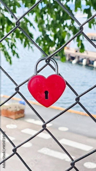 Fototapeta A heart-shaped padlock attached to a chain link fence with an urban cityscape visible in the background