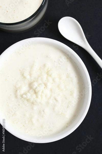 Fototapeta Kefir grains in milk with a glass of fresh kefir drink on the side, photographed overhead on slate with natural light (Selective Focus, Focus on the top of the kefir grains and the kefir drink)