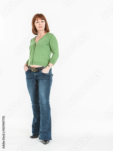 Fototapeta Unimpressed. A neutral yet neutral look on the face of a middle aged woman standing with her hands in her pockets with direct eye contact to the camera. Full length studio portrait isolated on white. 